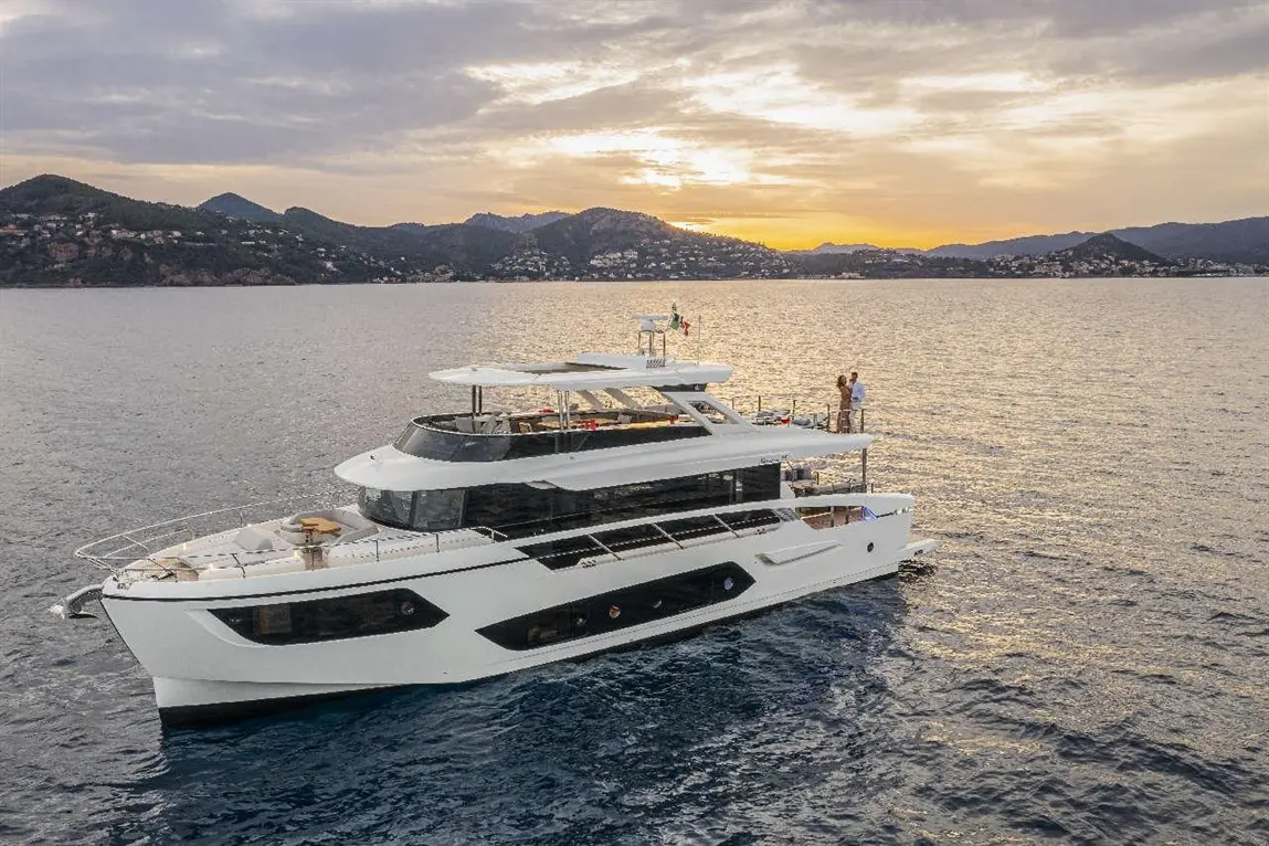 Luxury yacht at sunset with people on deck, surrounded by calm waters and mountainous coastline in the background.